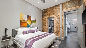 A modern hotel room with a large bed, a brick accent wall, and a view into the hallway. - Poolside 3BR w Hot Tubs, Grill Near French Qtr, Great for Families & Groups (New Orleans)