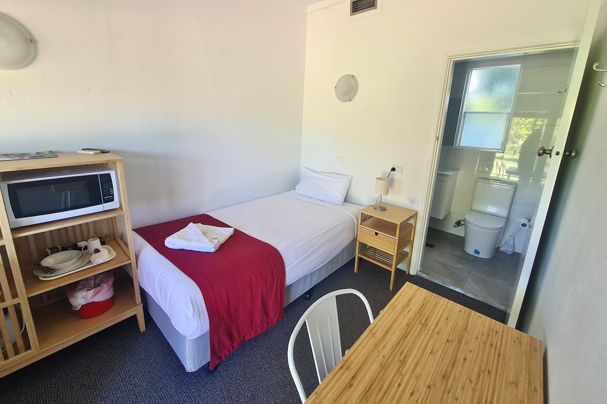 A single bed with a red blanket, a wooden nightstand with a lamp, a microwave on a shelf, and a view of the bathroom through an open door.