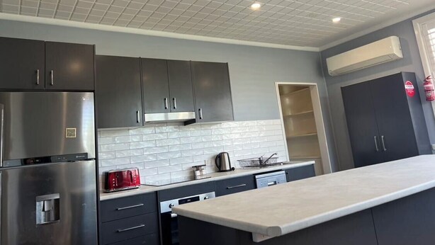 A modern kitchen with grey cabinets, a stainless steel refrigerator, a built-in oven, a white tiled backsplash, and a large island with a light-colored countertop. - Bridge Motel Wellington (Wellington)