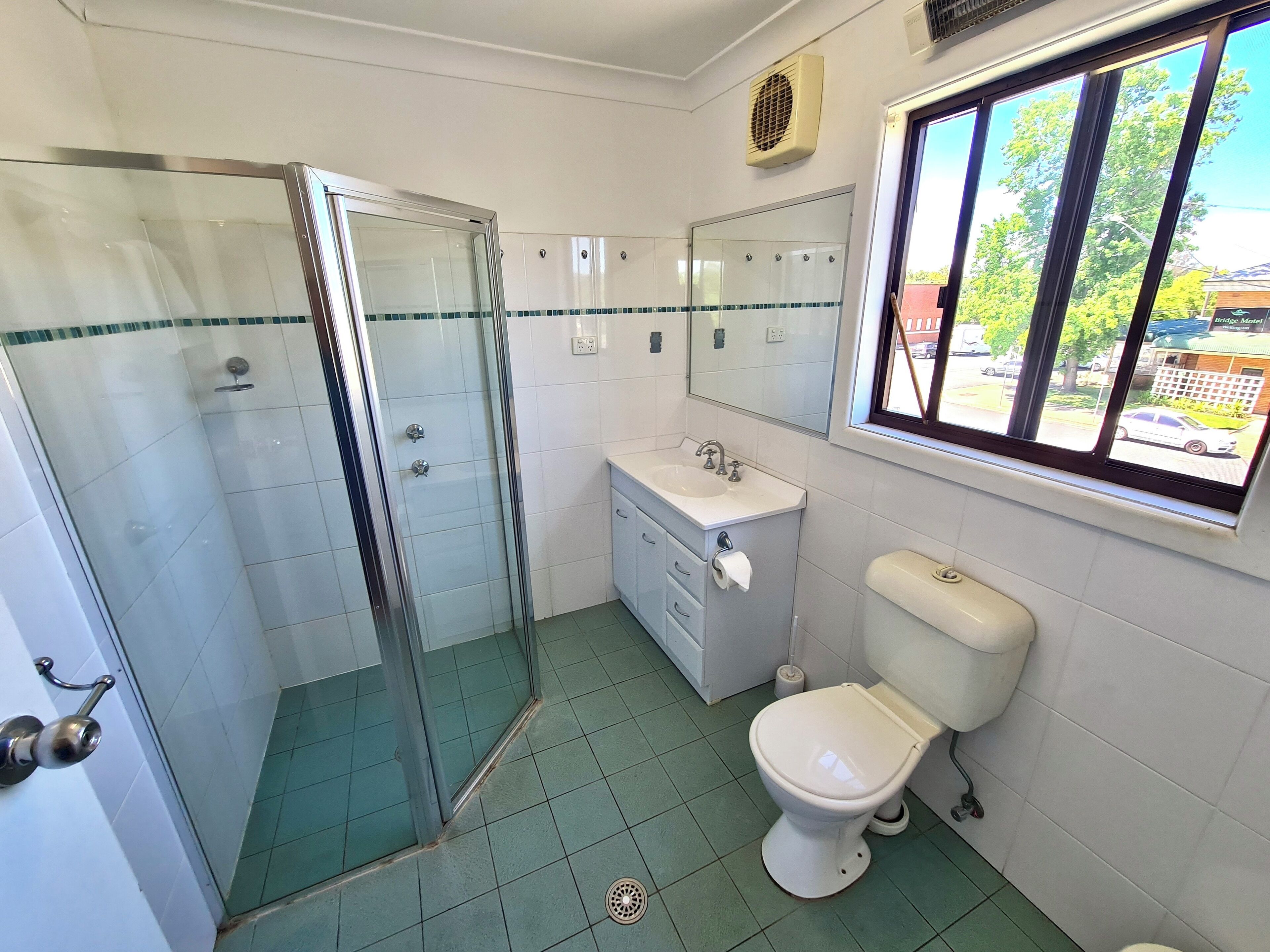 A bathroom with a glass shower enclosure, a sink, a toilet, and a window with a view of a motel outside.