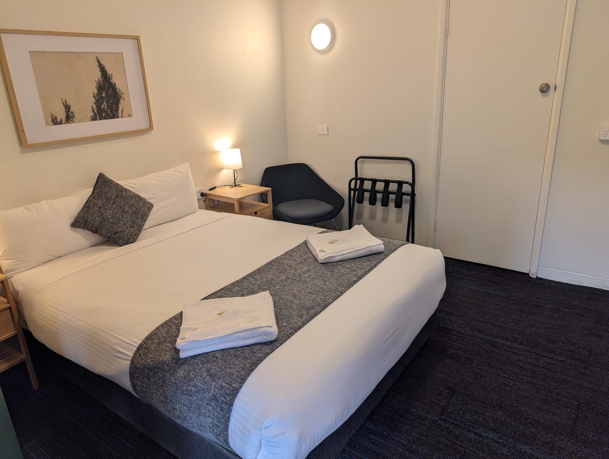 A hotel room with a bed, a nightstand, a chair, a wall-mounted towel rack, and a framed picture on the wall.