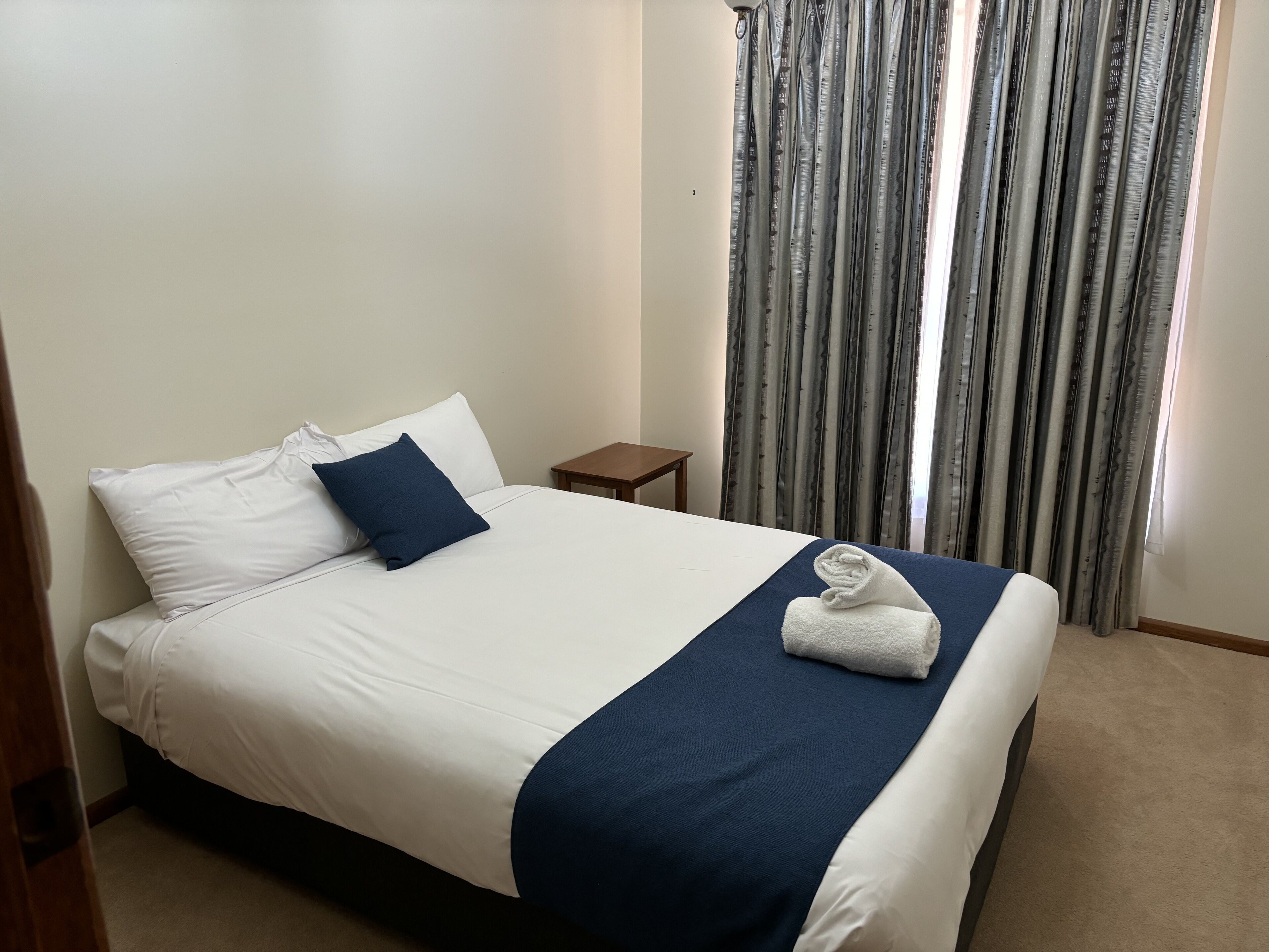 Un letto rifatto con cura, lenzuola bianche e blu navy, un piccolo comodino in legno e una finestra con tende a fantasia.