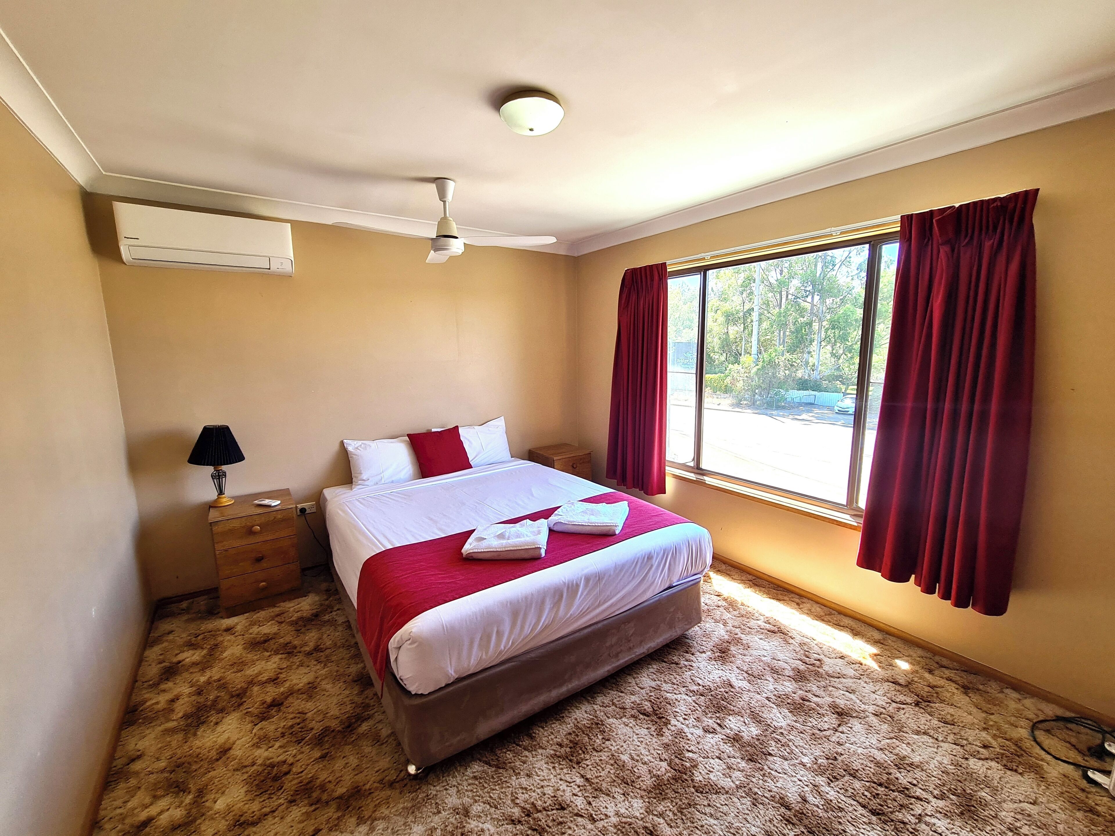 A bedroom with a bed, a ceiling fan, an air conditioning unit, a nightstand, a lamp, and a window with curtains.