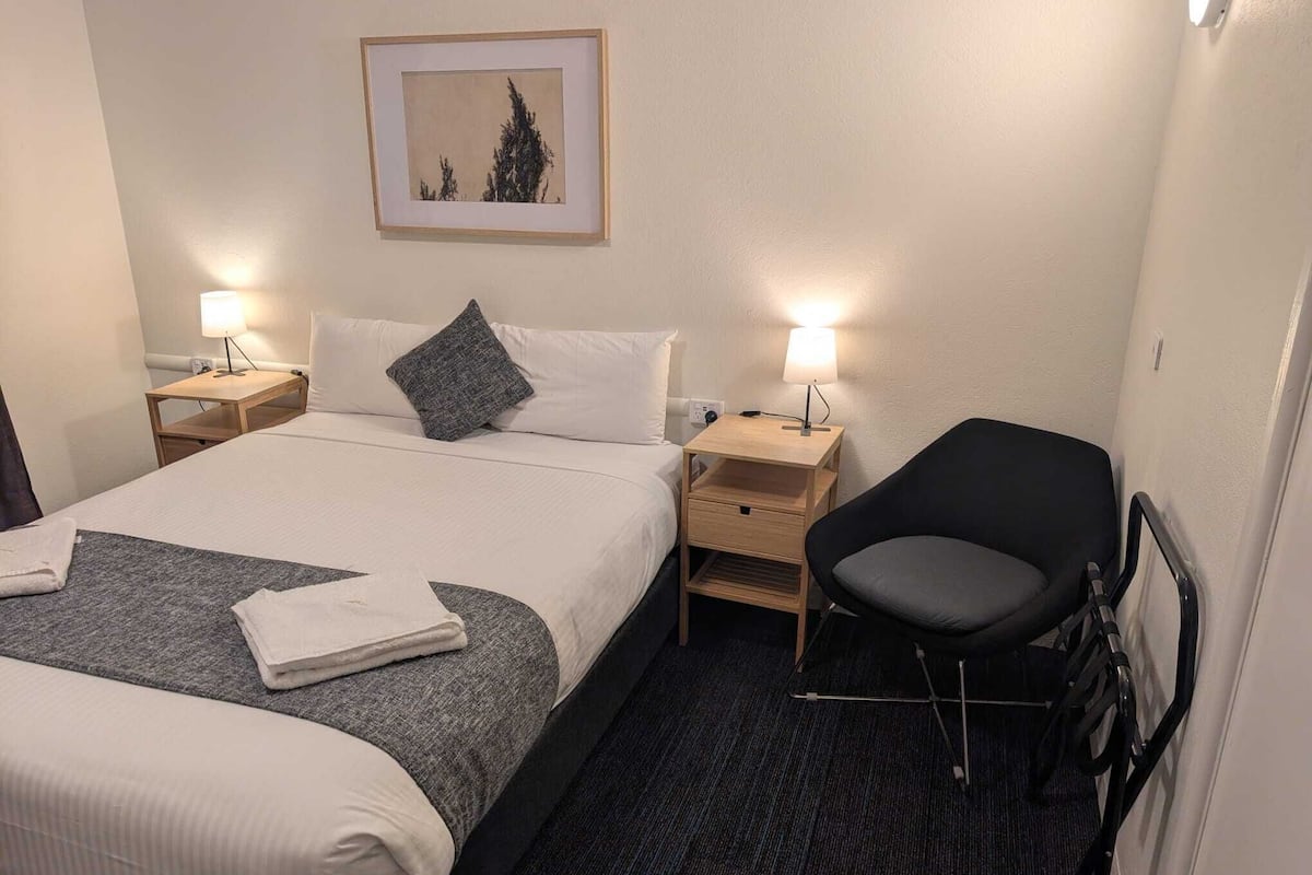 A hotel room with a bed, two bedside tables, a chair, a framed picture on the wall, and a carpeted floor.