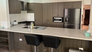 A modern kitchen with dark cabinets, a stainless steel refrigerator, a microwave, and a gas stove. There are two black bar stools and a small sink with a dish soap bottle. - Bridge Motel Wellington (Wellington)
