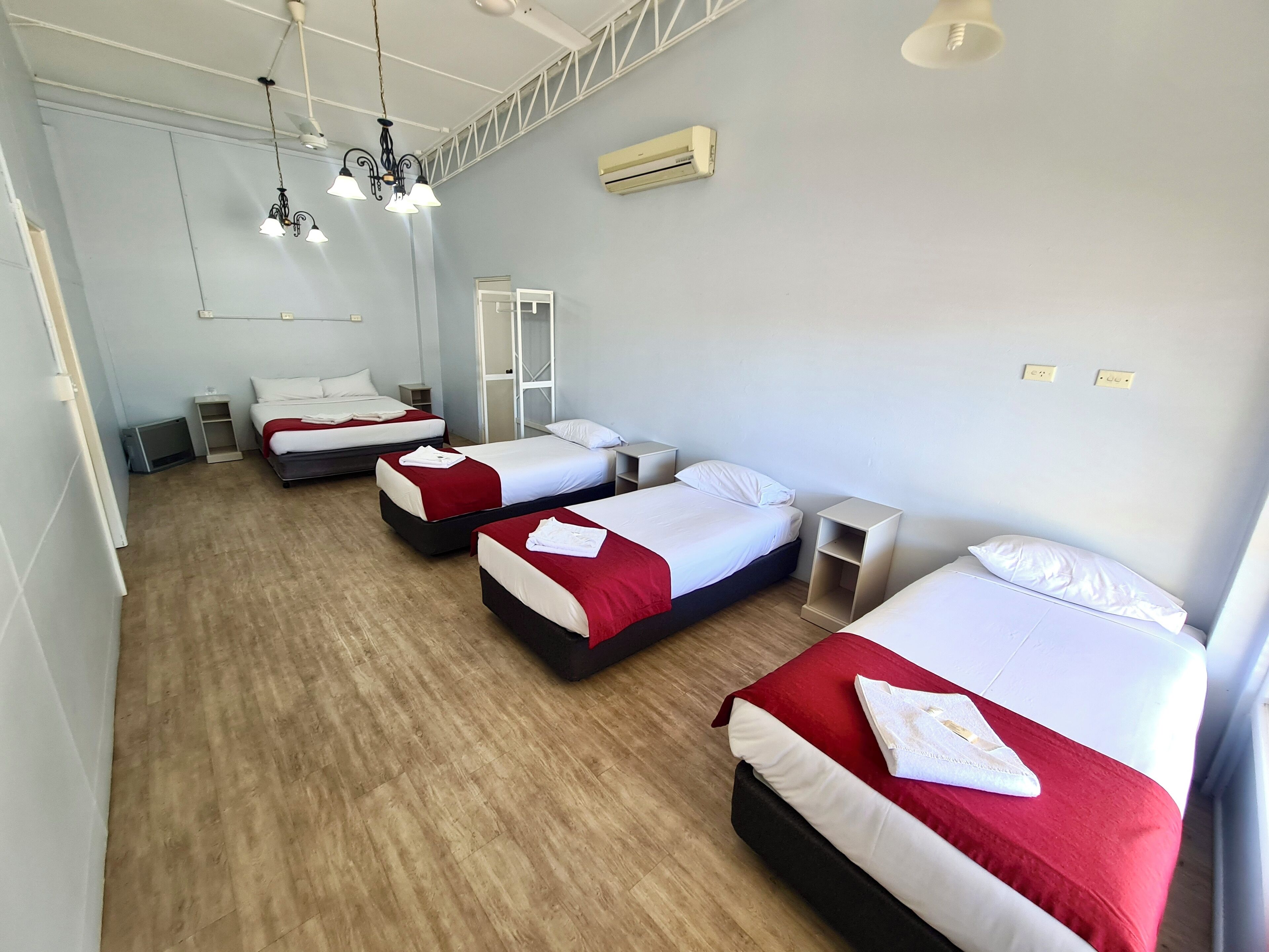 A hotel room with three beds, each with white and red bedding, a wooden floor, and a ceiling fan.