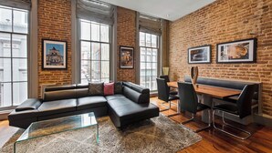 A modern interior with a brick wall, large windows, a sectional sofa, a dining table with chairs, and artwork on the walls.