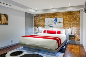 A modern bedroom with a brick wall, a large bed with red and white bedding, a cityscape painting, and a nightstand with a lamp. - Private Hot Tub, Patio, & Grill! Heated Pool in Courtyard, Family Friendly (New Orleans)