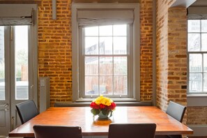 A dining area with a brick wall, a large window, and a wooden table with chairs. - Private Hot Tub, Patio, & Grill! Heated Pool in Courtyard, Family Friendly (New Orleans)