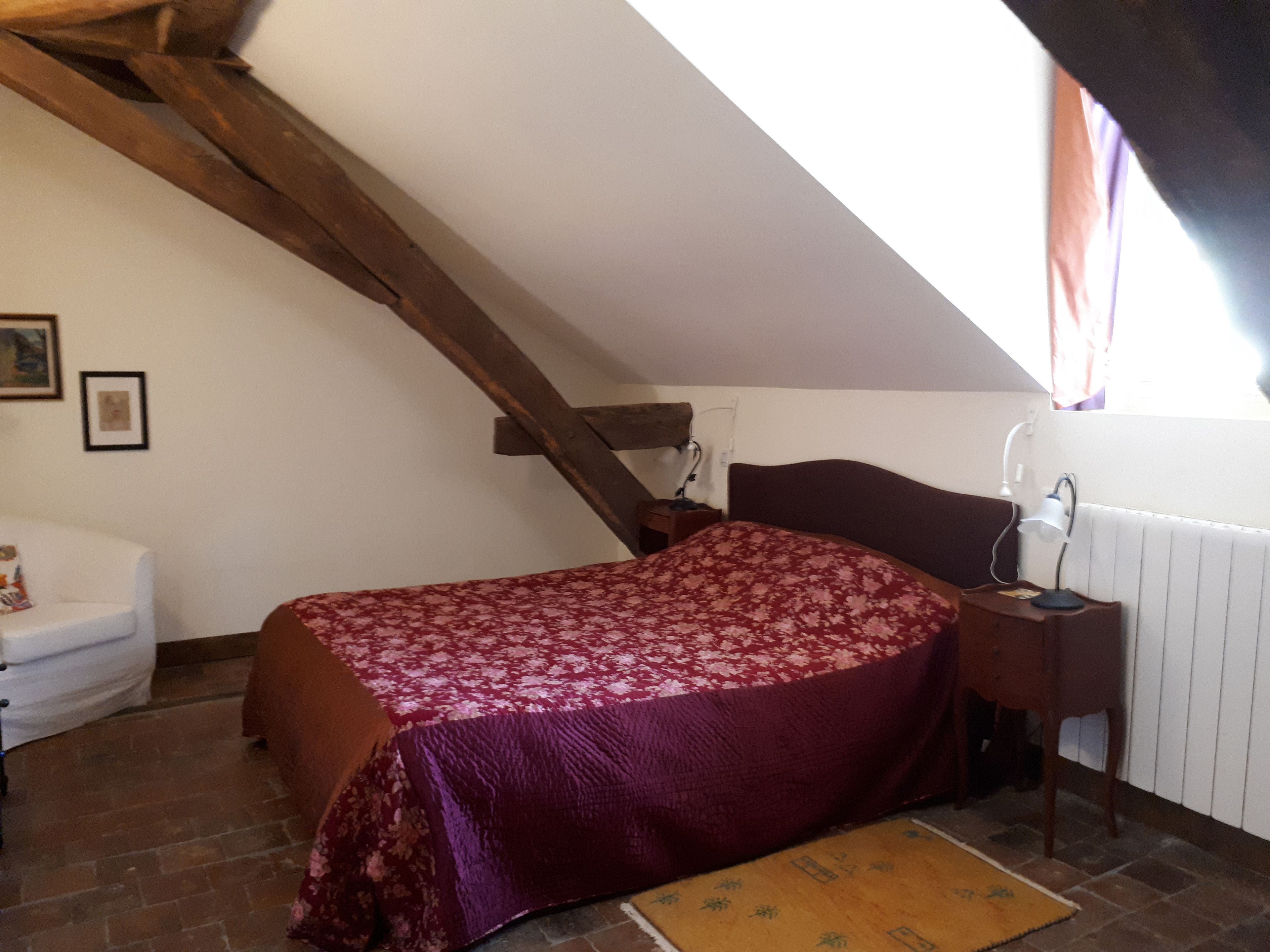Un dormitorio con techo inclinado, cama con colcha floral, mesita de noche con lámpara, una silla y un radiador.