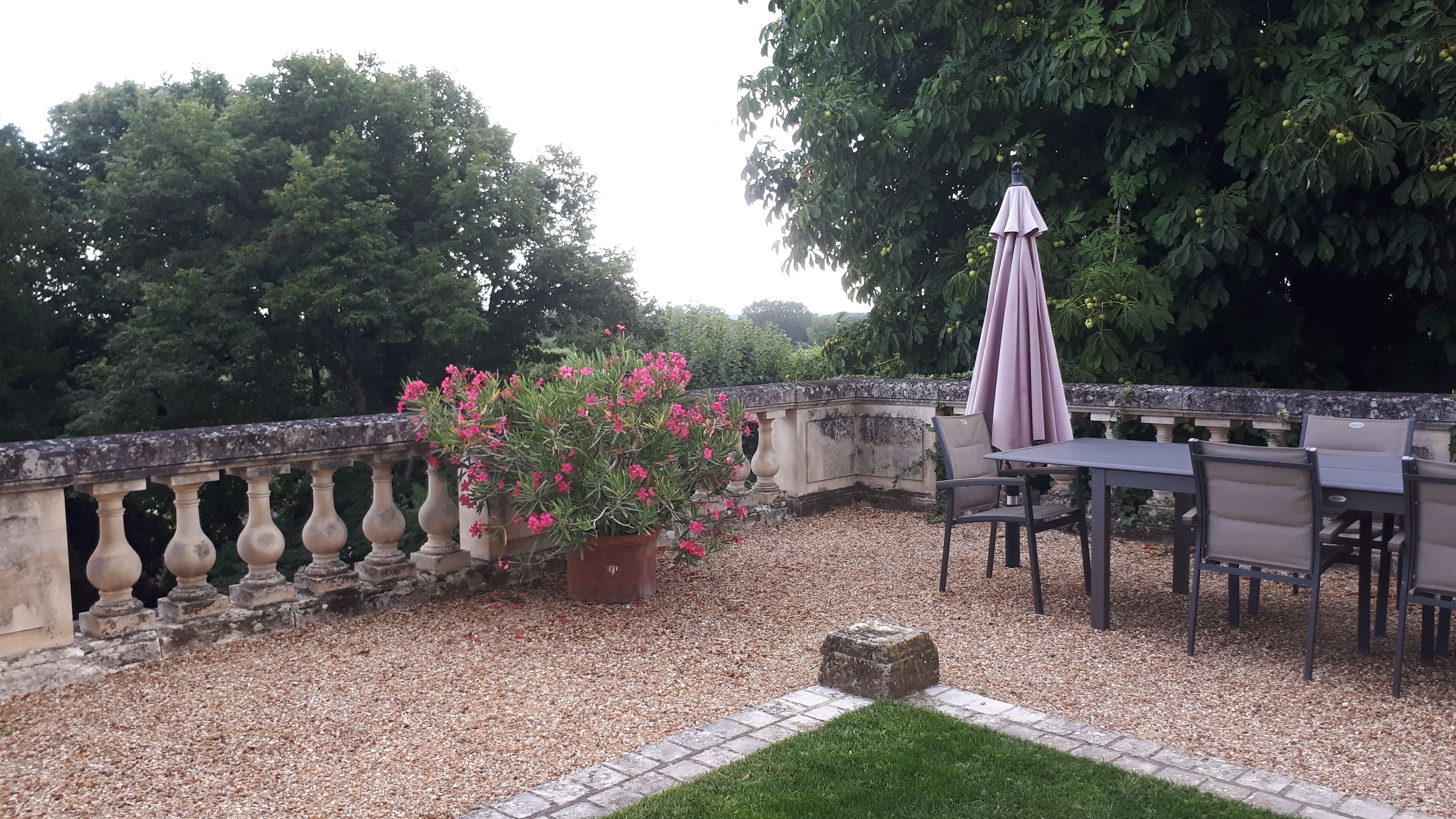 An outdoor patio with a table and chairs, surrounded by a stone balustrade and a potted plant with pink flowers.
