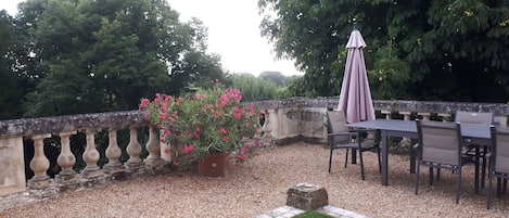 An outdoor patio with a table and chairs, surrounded by a stone balustrade and a potted plant with pink flowers.