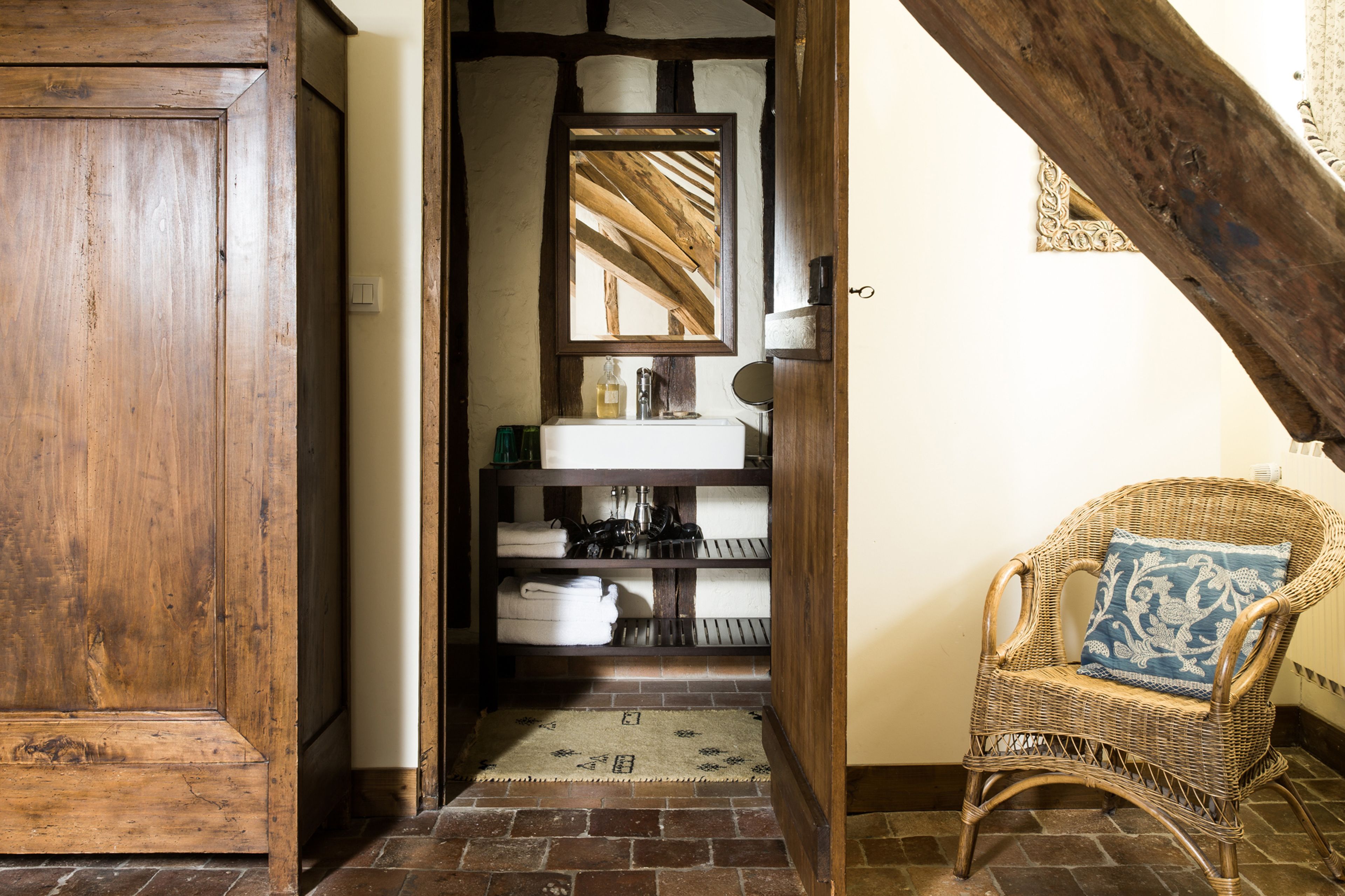 A rustic bathroom with a wooden door, a white sink, a mirror, and a wicker chair with a cushion.