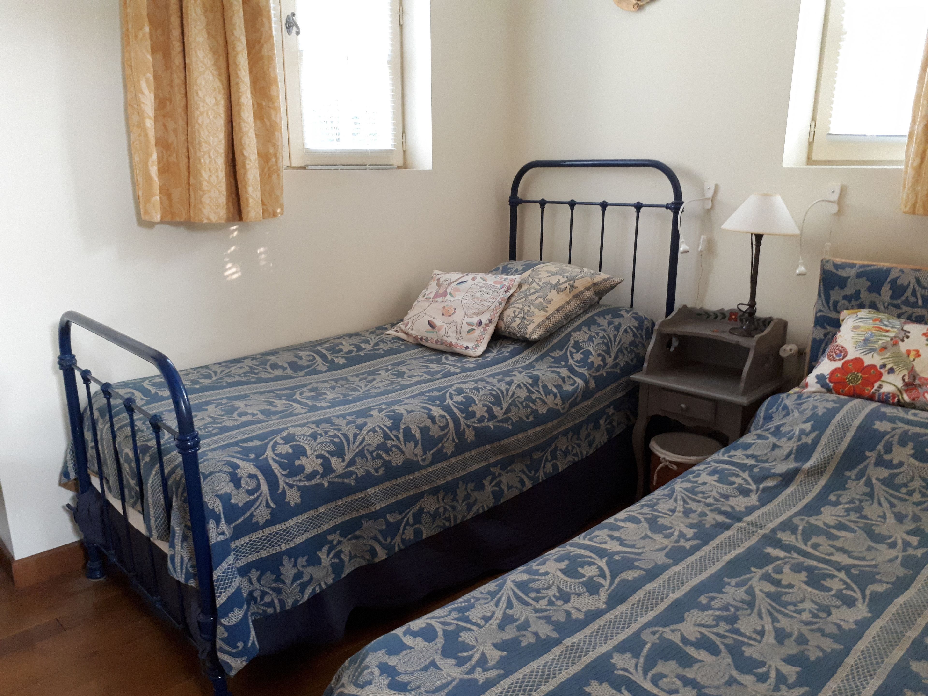 A small, single-bed room with a metal bed frame, a bedside table with a lamp, and a window with curtains.