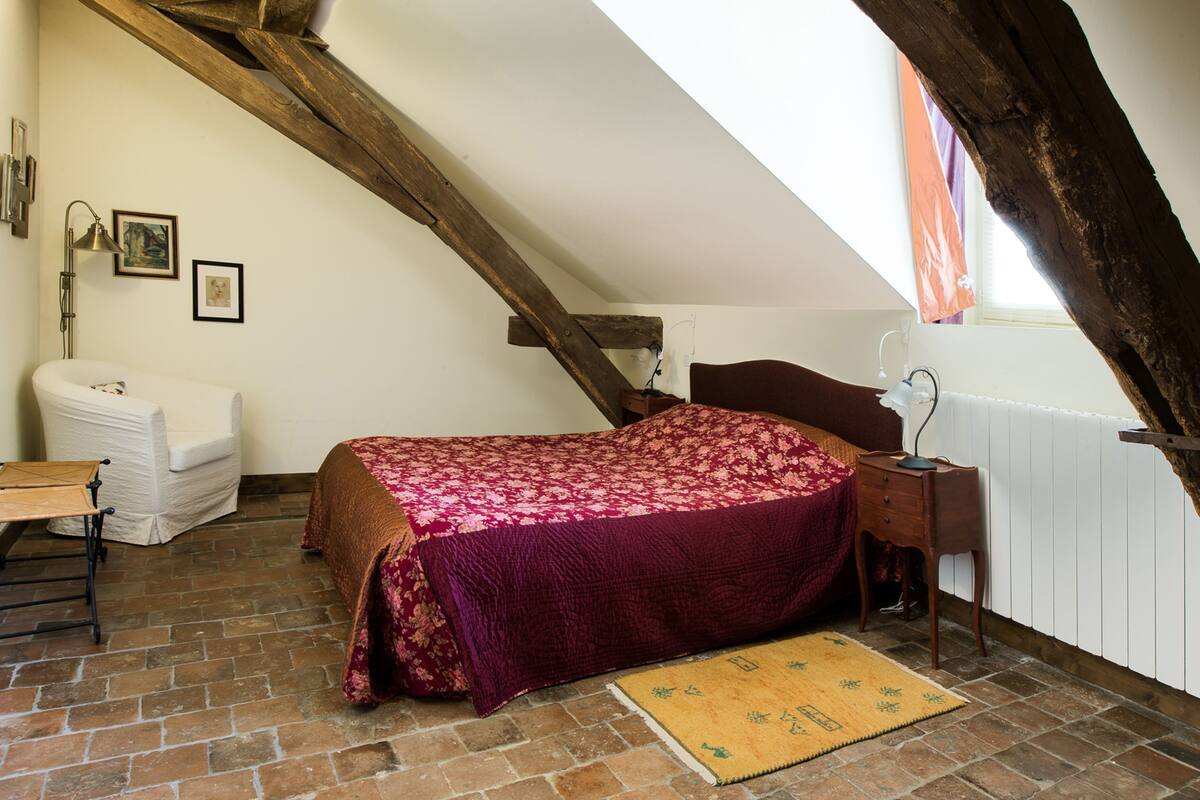 A loft bedroom with a sloped ceiling, a bed with a floral bedspread, a white armchair, a wooden nightstand, and a radiator.