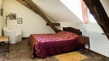 A loft bedroom with a sloped ceiling, a bed with a floral bedspread, a white armchair, a wooden nightstand, and a radiator.