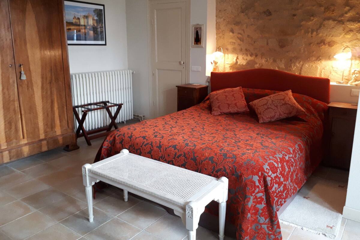 A bedroom with a bed, a wooden wardrobe, a radiator, a white bench, and a stone wall.