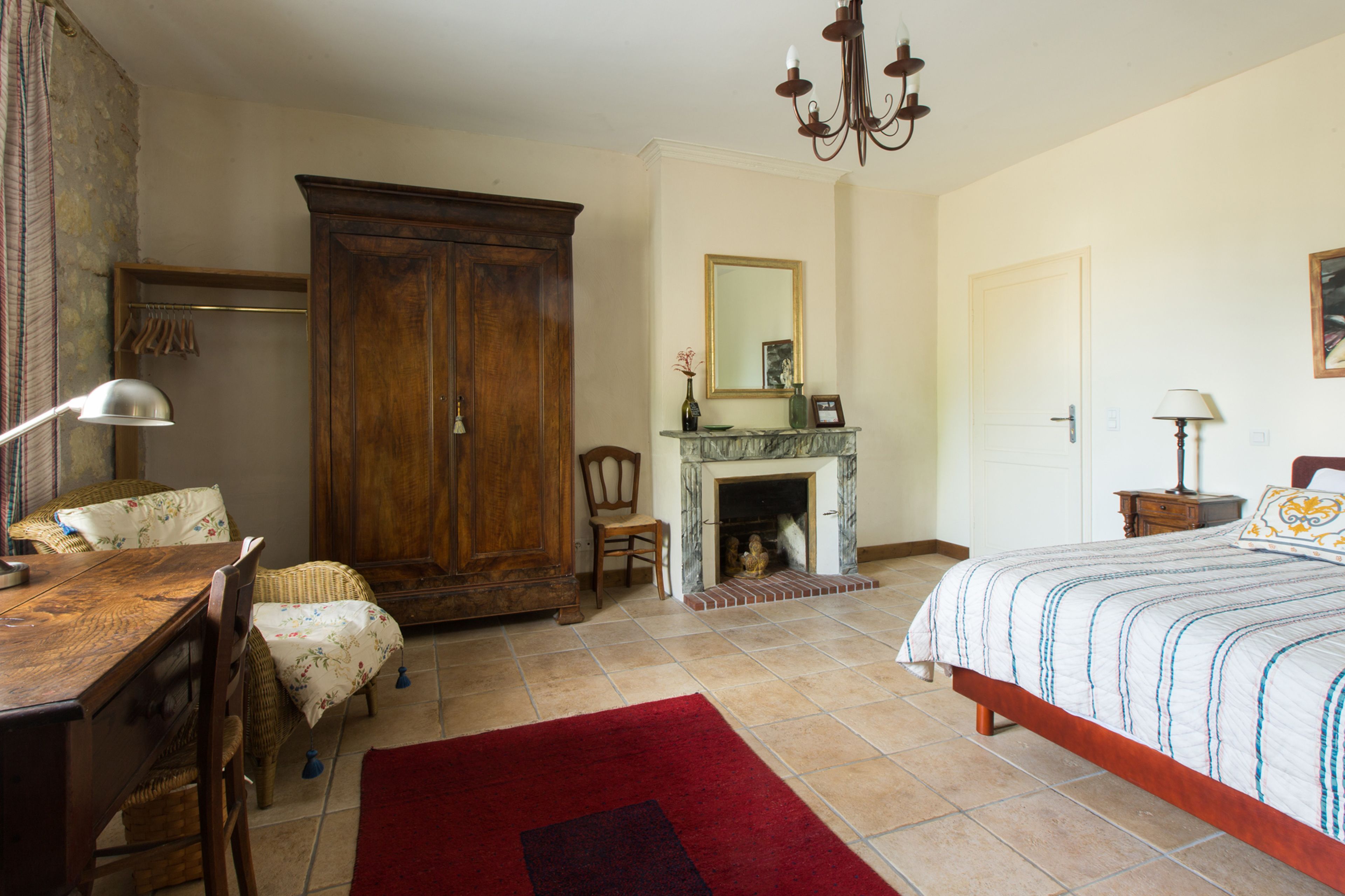Un dormitorio con una cama de madera, una chimenea, un armario grande de madera, una mesa de comedor con sillas y una alfombra roja.