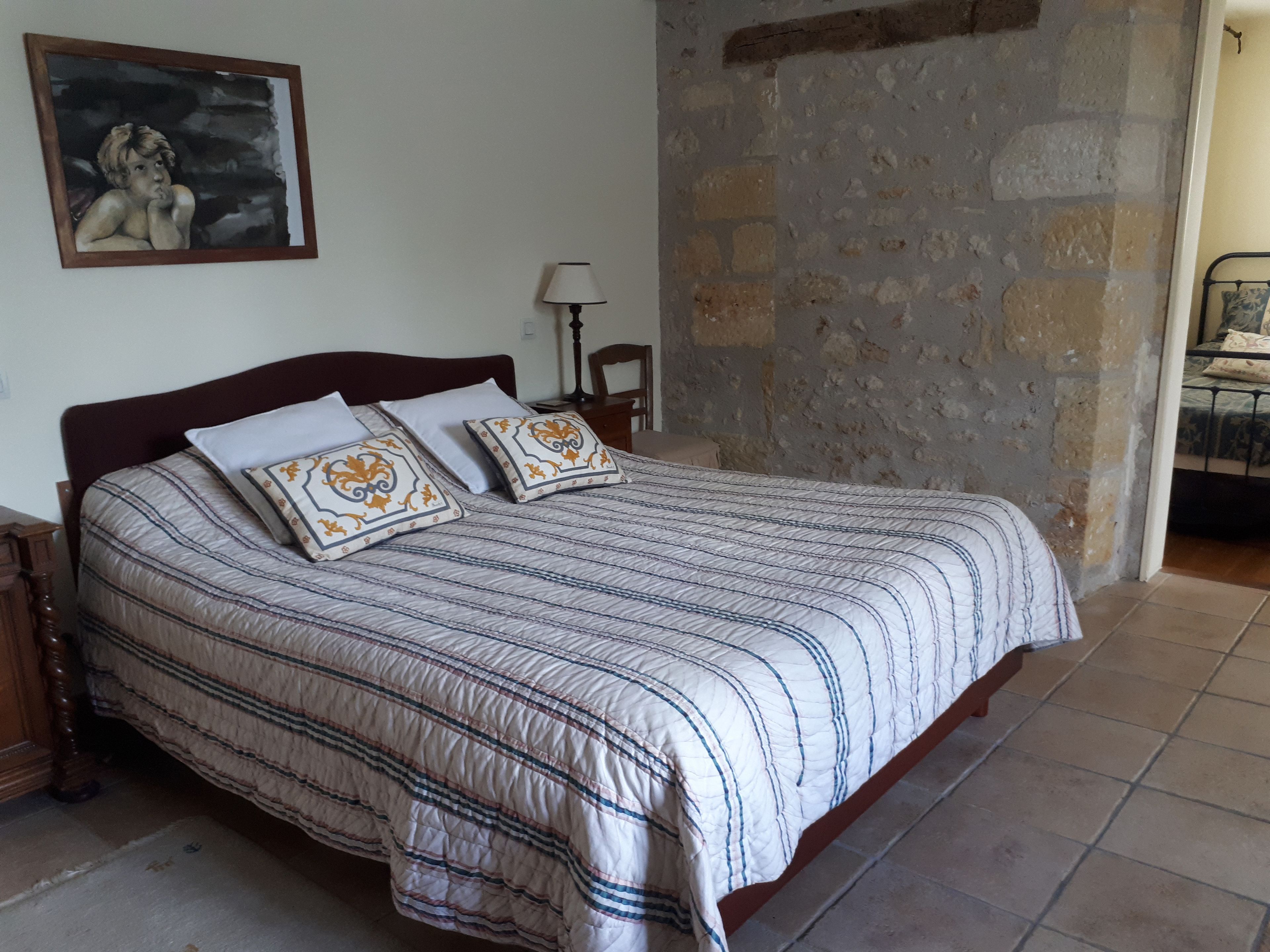 A bedroom with a large bed, a stone wall, a painting on the wall, and a nightstand with a lamp.