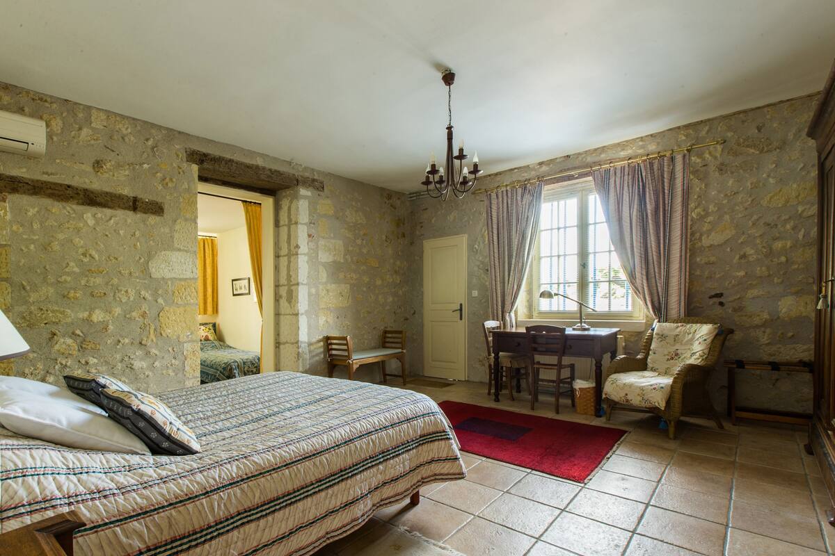 A bedroom with a bed, a desk, and a chair. There is a window with curtains, a chandelier, and a stone wall.