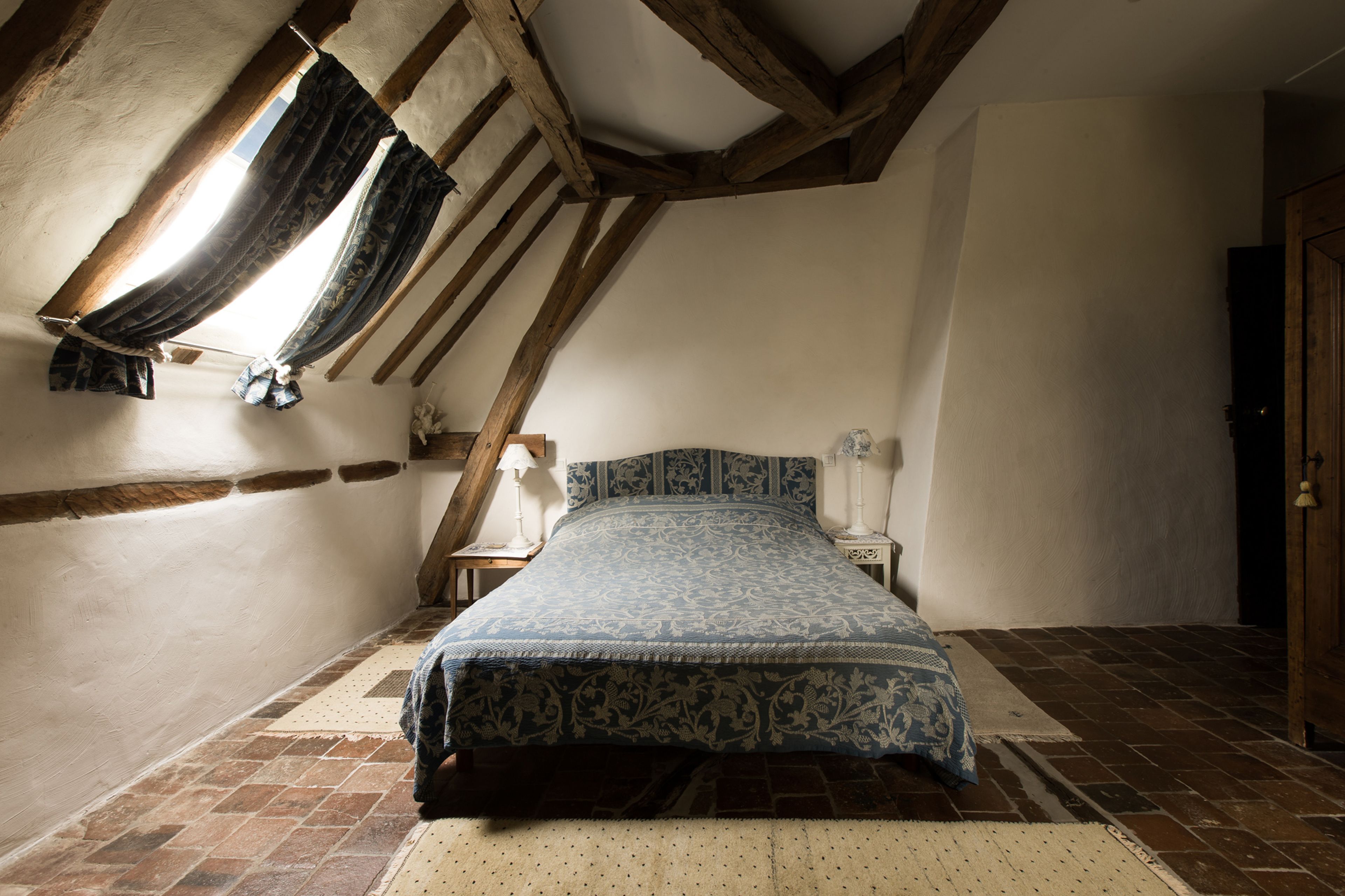 Ein Schlafzimmer mit geneigter Decke, einem Bett mit gemustertem Überwurf, einem Nachttisch mit Lampe und einem Fenster mit Vorhängen.