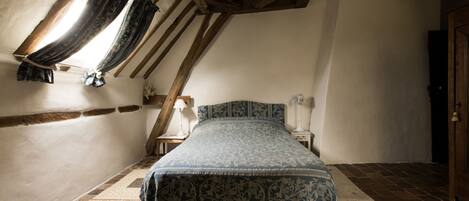 Ein Schlafzimmer mit geneigter Decke, einem Bett mit gemustertem Überwurf, einem Nachttisch mit Lampe und einem Fenster mit Vorhängen.
