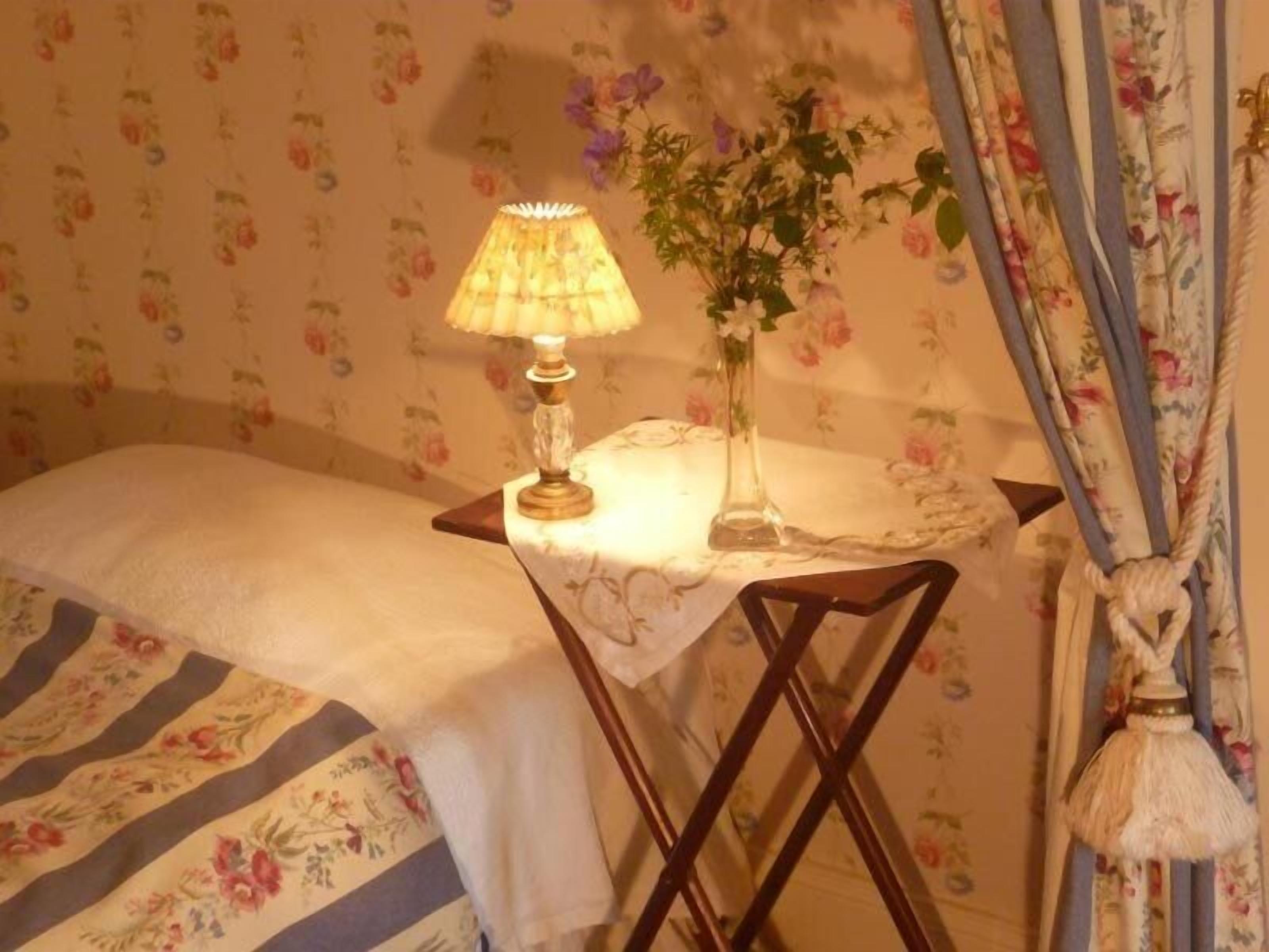 A bedroom with a bed, a bedside table with a lamp, a vase with flowers, and floral curtains.
