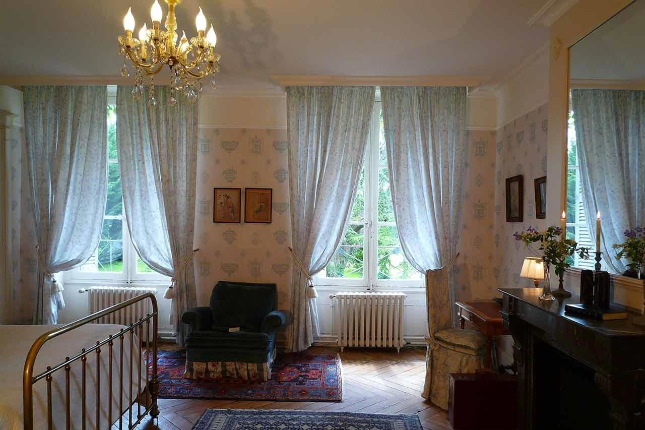 Une pièce avec un lustre, deux fenêtres avec des rideaux, une chaise, un radiateur et une cheminée.