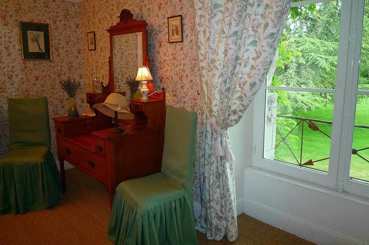 A room with floral wallpaper, a wooden dresser, two green chairs, a mirror, and a window with a view of greenery.