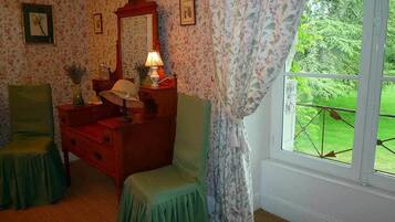 A room with floral wallpaper, a wooden dresser, two green chairs, a mirror, and a window with a view of greenery.