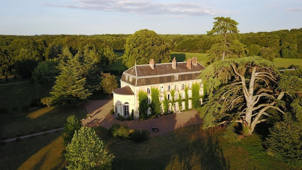 Een groot, wit, twee verdiepingen tellend gebouw met een symmetrische gevel, omgeven door weelderige beplanting en bomen.