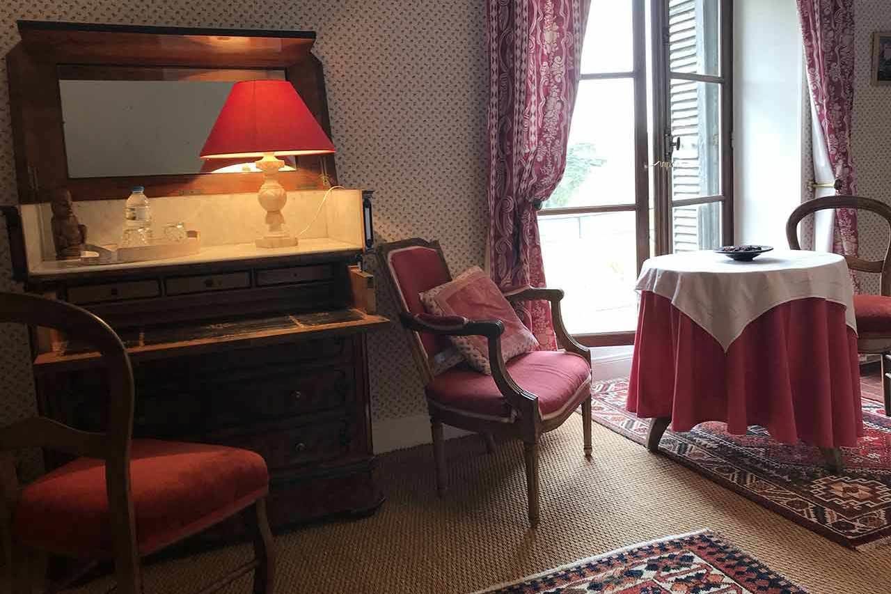 A cozy room with a wooden dresser, a red chair, a table with a white tablecloth, and a window with curtains.