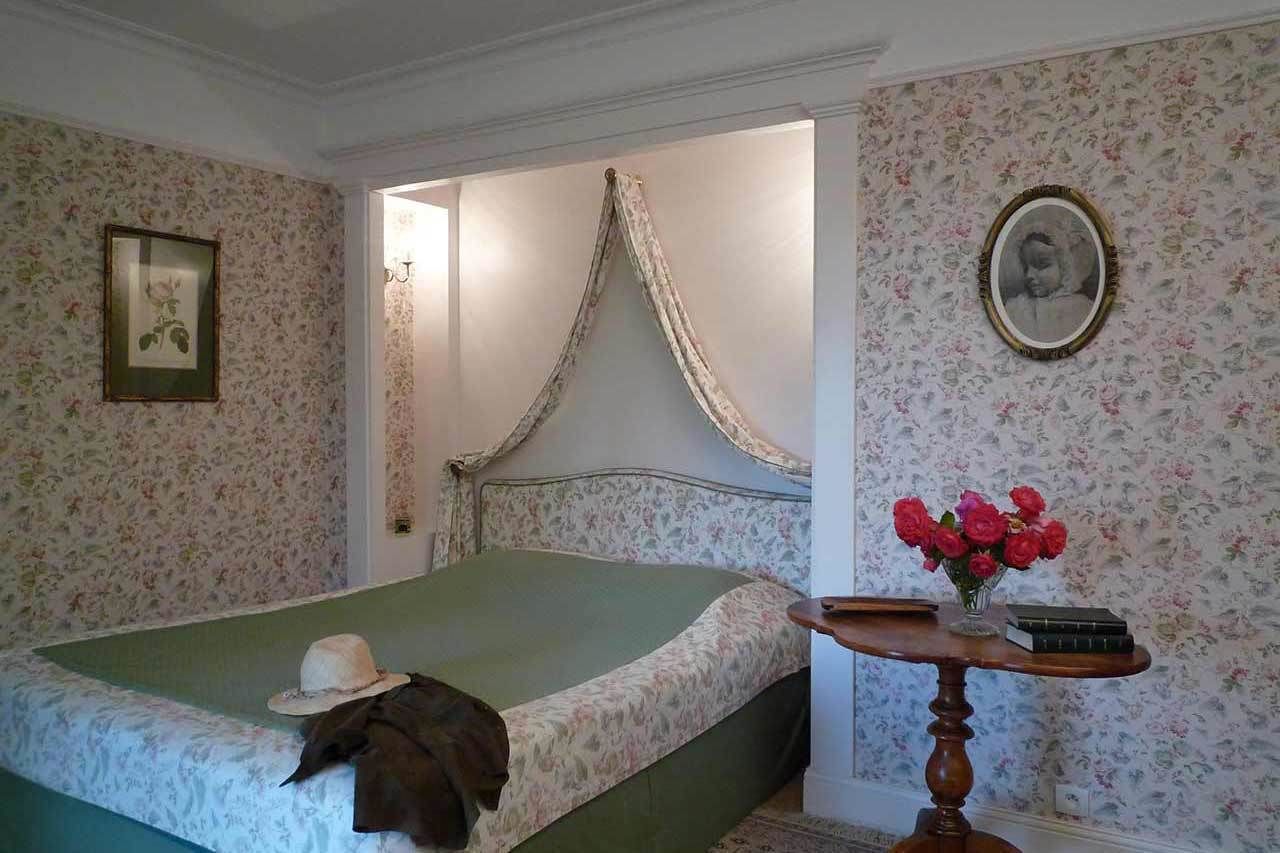 Een slaapkamer met een bed, een nachtkastje met een boek en bloemen, een ingelijst beeld aan de muur, en een behang met bloemen.