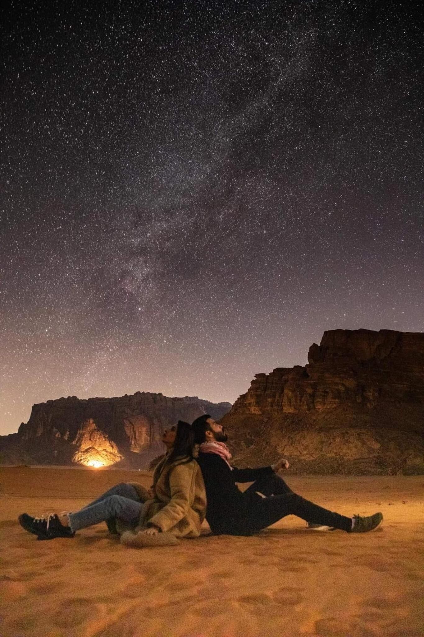 Una pareja sentada sobre la arena bajo un cielo estrellado con una montaña al fondo.