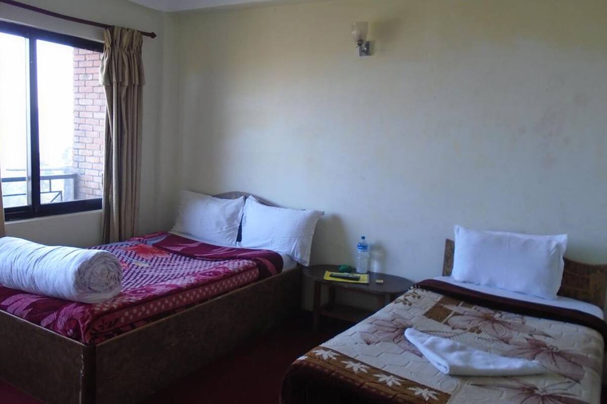 Habitación de hotel con dos camas, una mesita pequeña, un ventanal con cortinas y una pared de ladrillo.