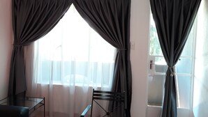 Ein Zimmer mit zwei schwarzen Vorhängen, einem Glastisch, einem Stuhl und einem Fenster mit Blick auf Bäume.