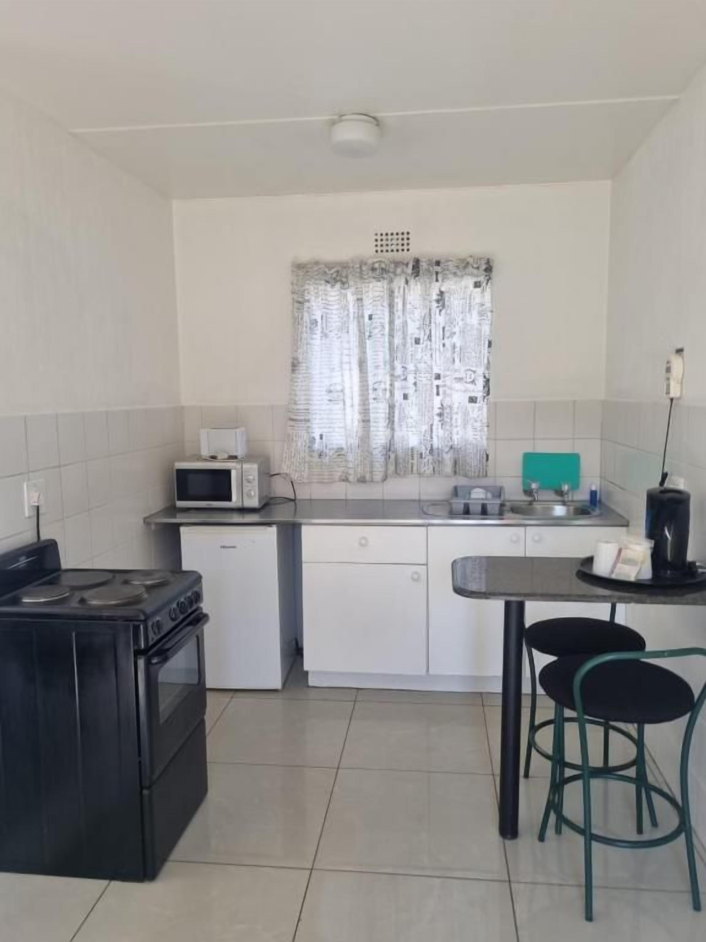 A compact kitchen with a microwave, toaster, and sink. There is a small refrigerator and a stove. The room has white walls and a window with curtains.
