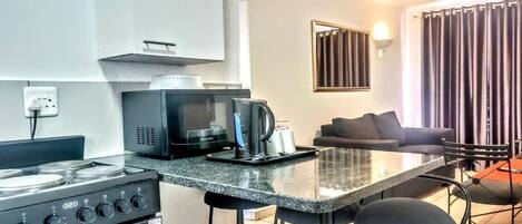 Cocina compacta con estufa a gas, microondas y cafetera. En la zona de estar, hay un taburete de bar y un sofá.