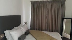 Ein Hotelzimmer mit Bett, Kissen, Decke, Kopfteil, einem Fenster mit Vorhängen und einem Spiegel.