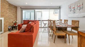 A modern living room with a red sofa, dining table with chairs, and a large window with blinds.