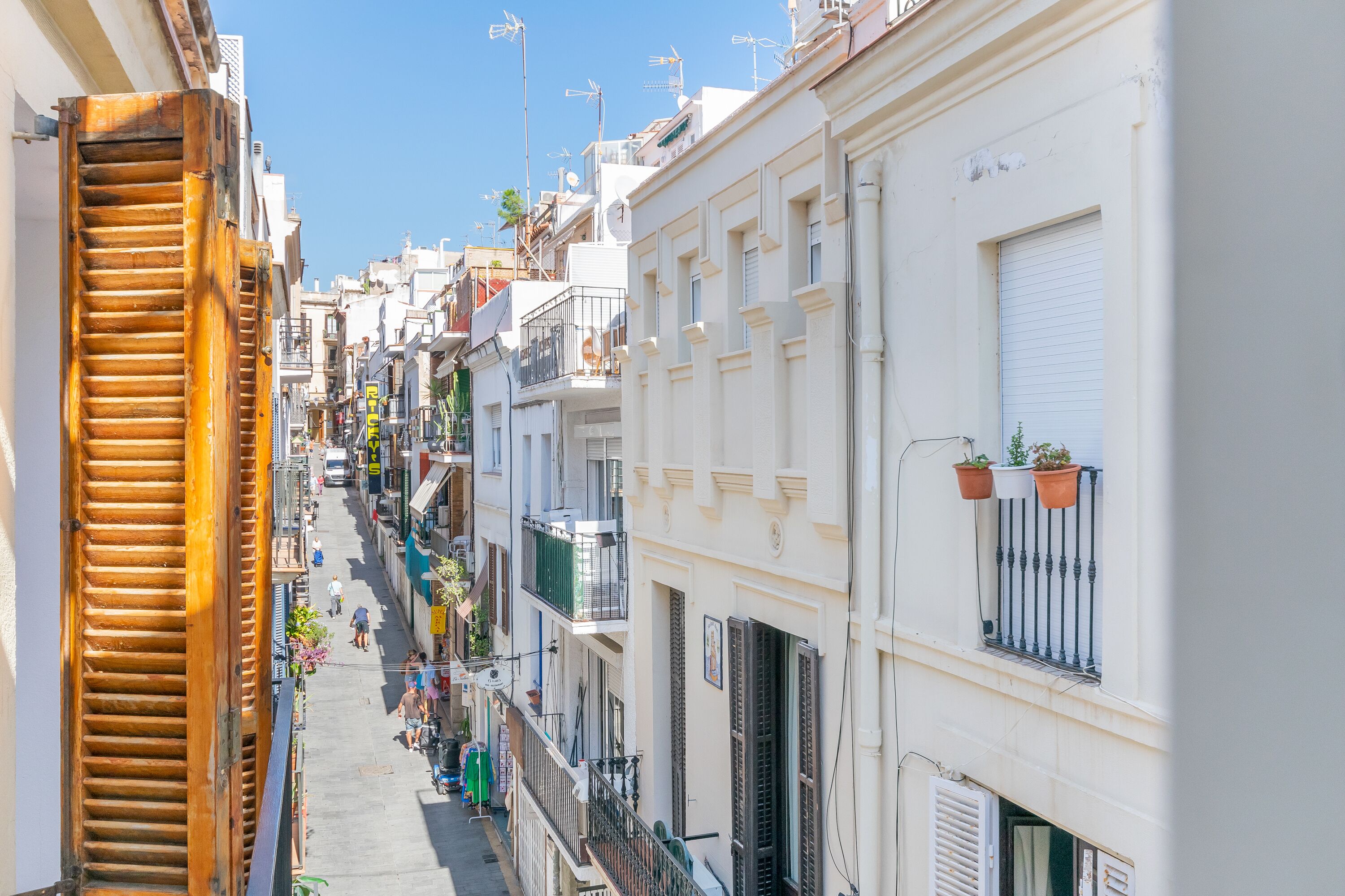 A narrow street lined with multi-story buildings, featuring balconies and potted plants.