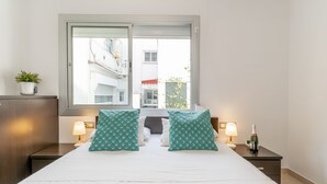 A hotel room with a large bed, two bedside lamps, a window with a view of buildings, and a small potted plant on the nightstand.