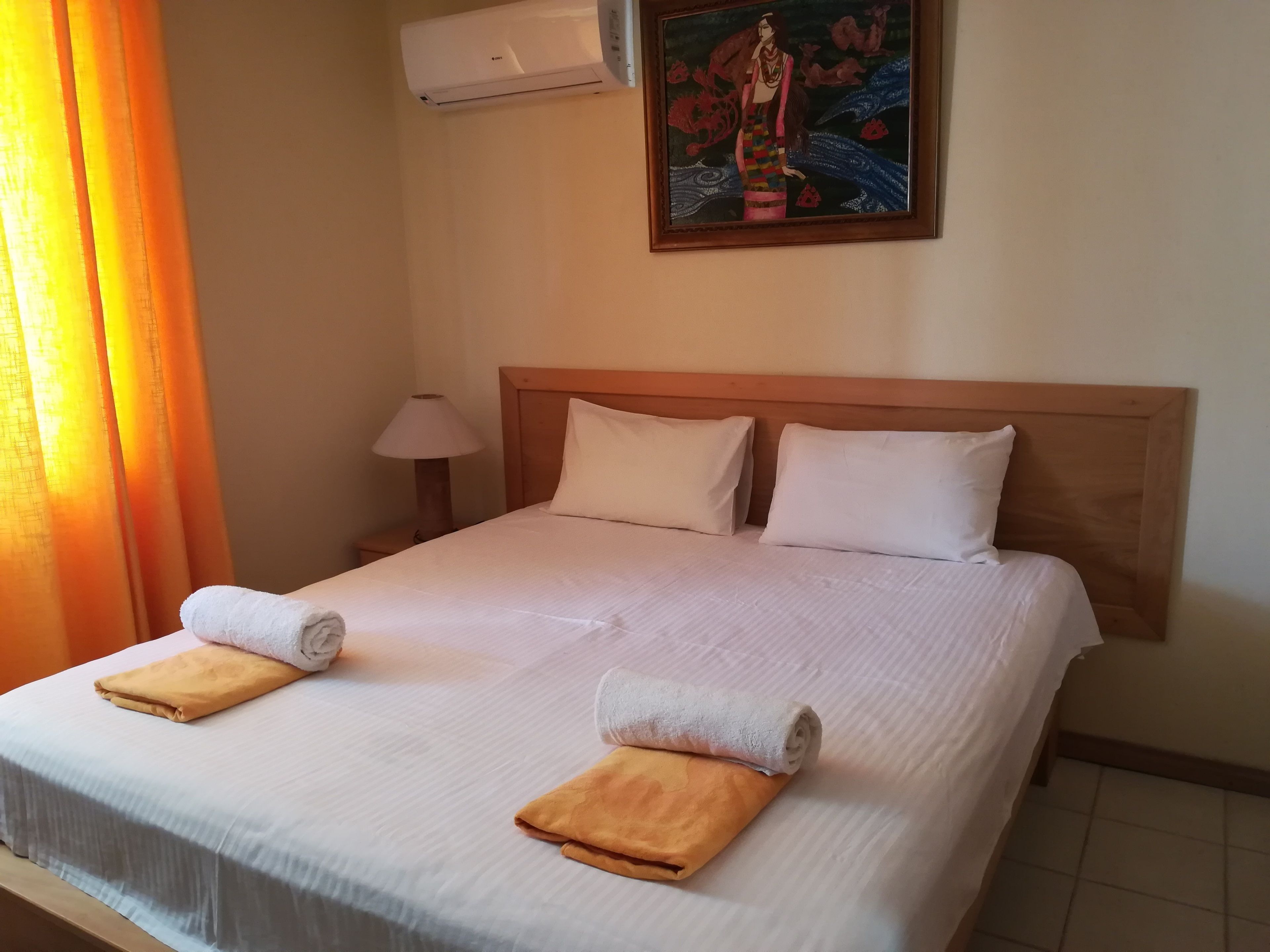 A neatly made bed with two pillows and two folded towels, an air conditioning unit above, and a painting on the wall.