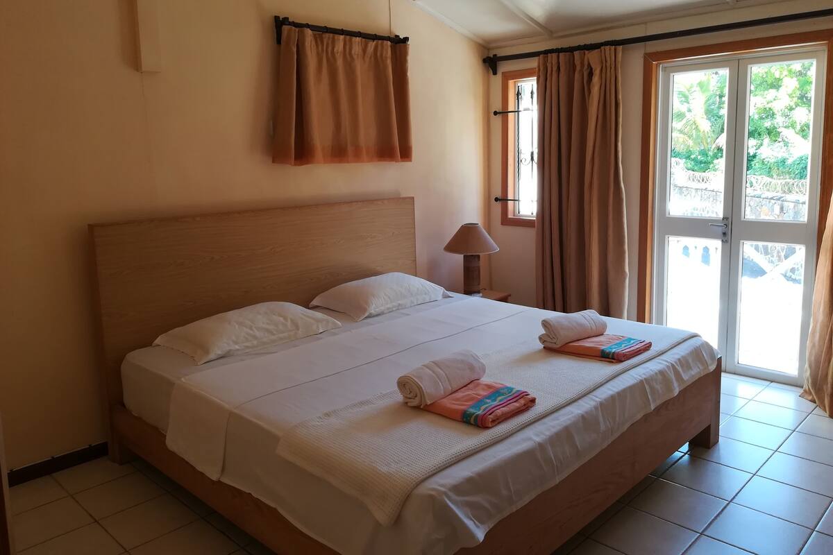 A hotel room with a large bed, a nightstand, a lamp, and a view of the outdoors through a window with curtains.