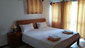 Cama bem arrumada com lençóis brancos, cabeceira de madeira e duas almofadas. Um criado-mudo com uma luminária e uma toalha dobrada. O quarto tem piso de azulejos e uma janela com cortinas.