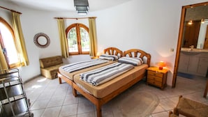 A bedroom with a double bed, a bedside table with a lamp, a wooden chair, a small sofa, and a cart.