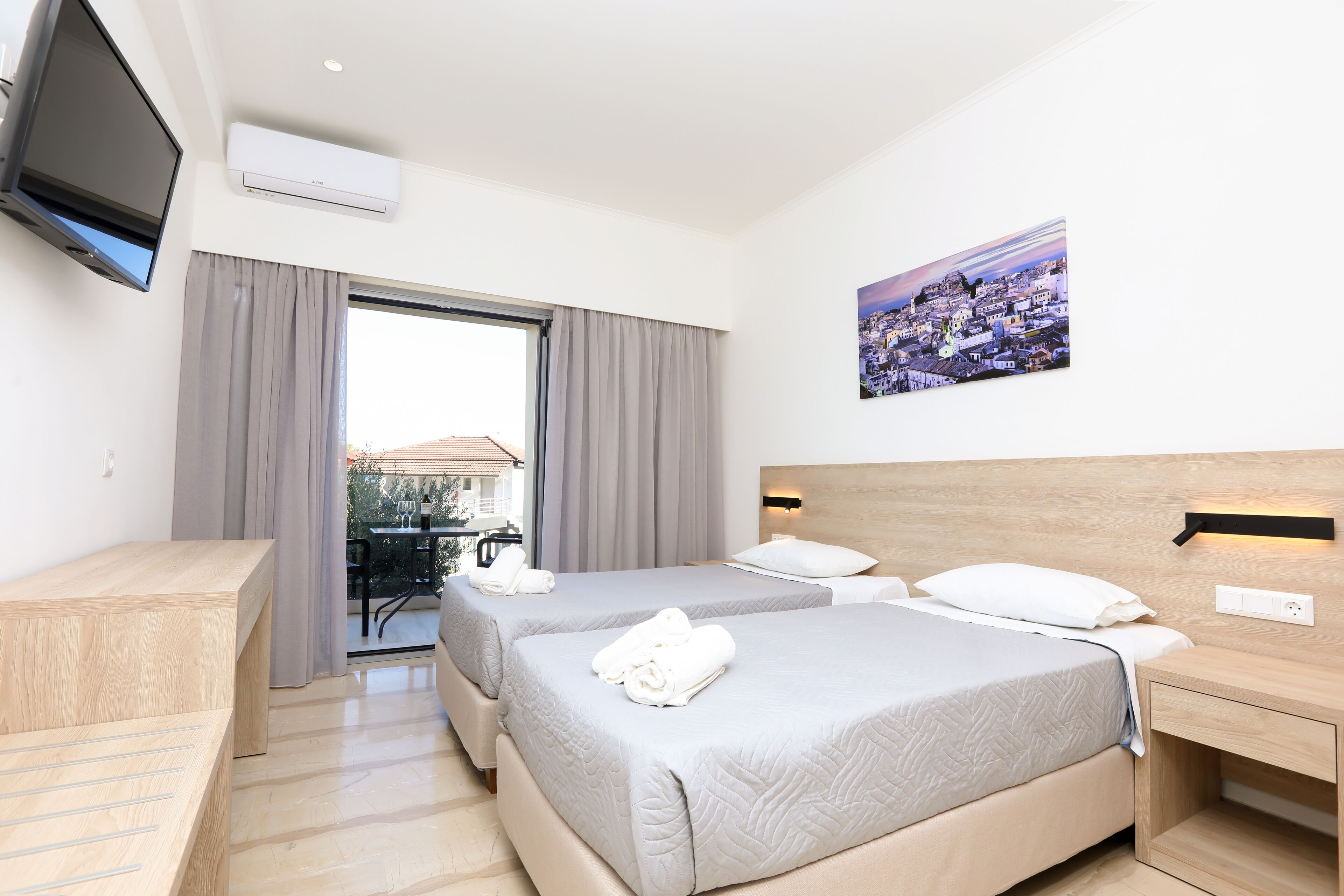 A hotel room with two beds, a TV, an air conditioner, a painting, and a view of the city.