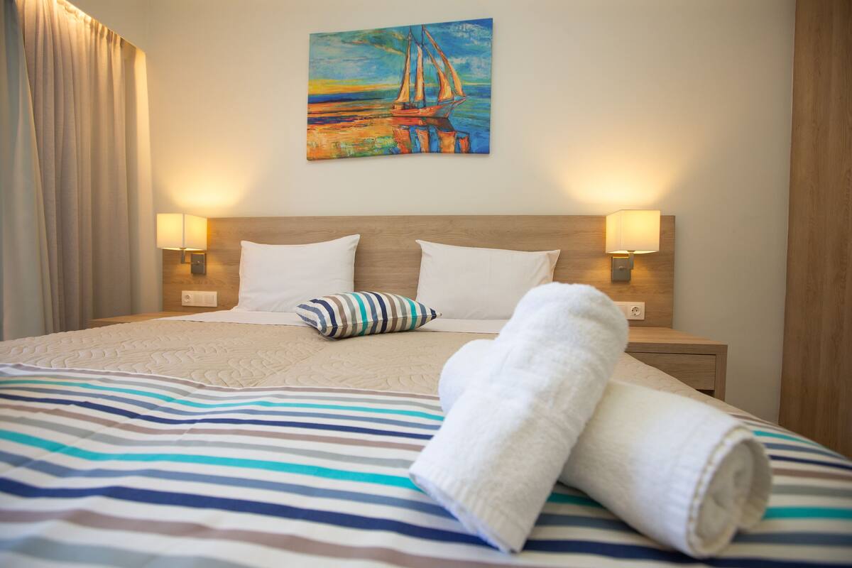A hotel room with a bed, striped bedding, a sailboat painting on the wall, two bedside lamps, and a wooden headboard.