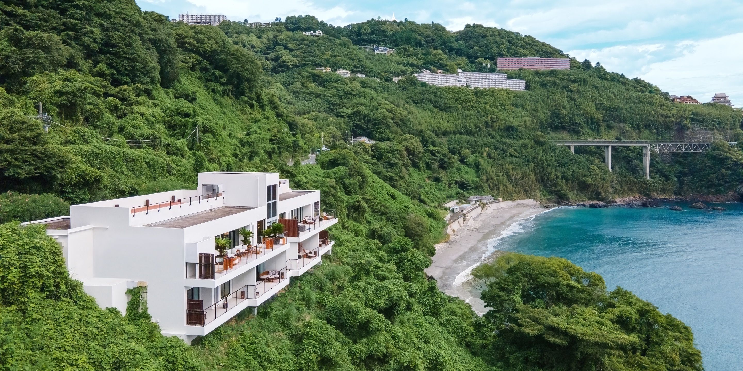 Un edificio moderno de varios pisos con balcones, ubicado en una colina con vista a una playa y un puente.