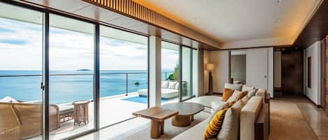 Suite de hotel moderna con un gran ventanal corredera que da a un balcón con vistas al mar. En el interior, hay un conjunto de sofás, una mesita y una lámpara de pie.